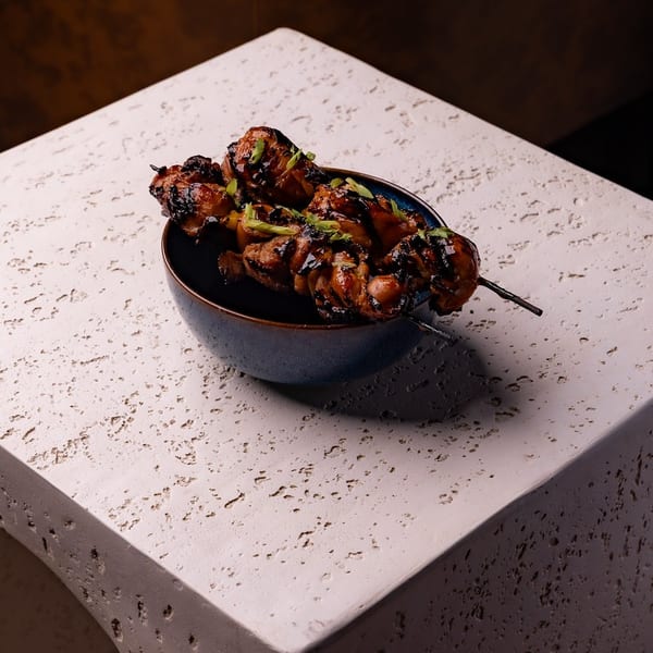 Glazed chicken skewers with green onions in a blue bowl on a textured stone block.