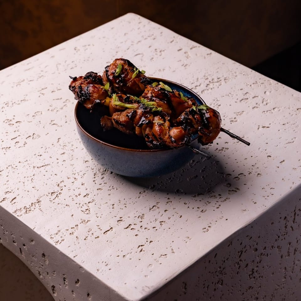 Glazed chicken skewers with green onions in a blue bowl on a textured stone block.