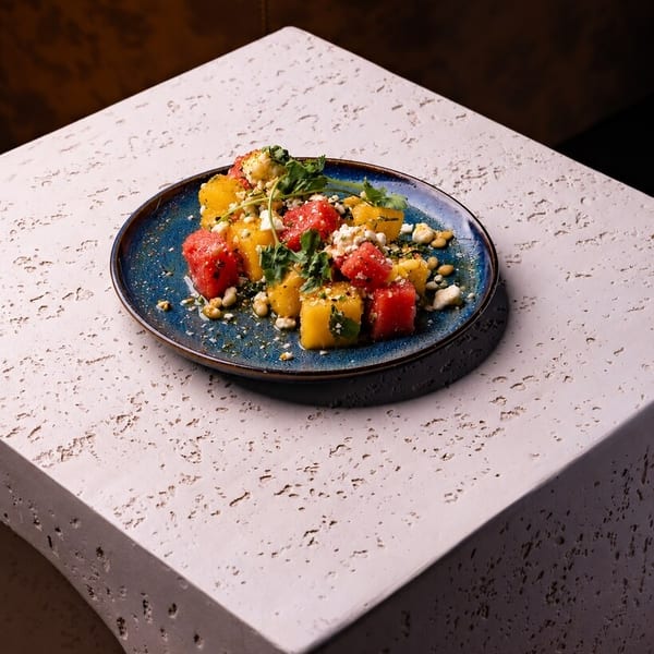 Vibrant watermelon and yellow fruit salad with feta, nuts, and greens on a blue plate.