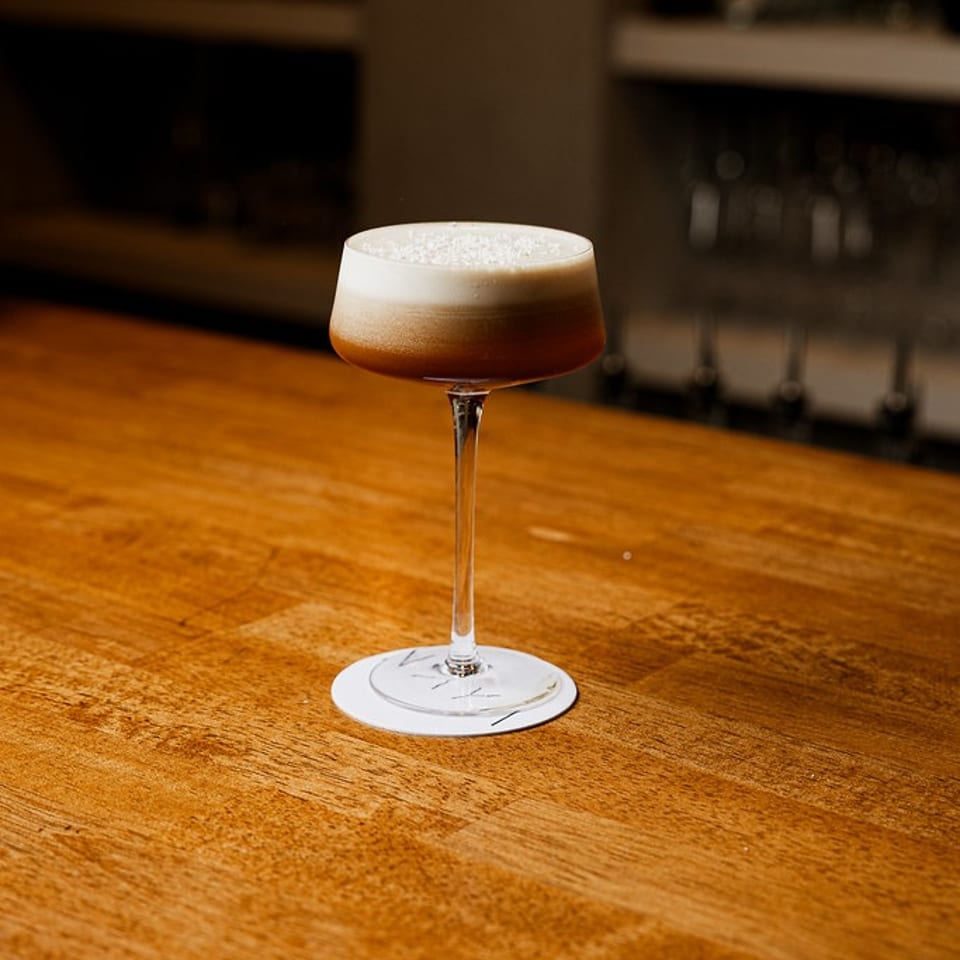 Dark cocktail with white foam in a stemmed glass on a wooden bar counter.