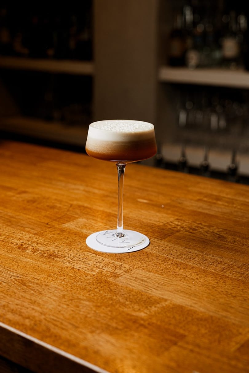 Dark cocktail with white foam in a stemmed glass on a wooden bar counter.