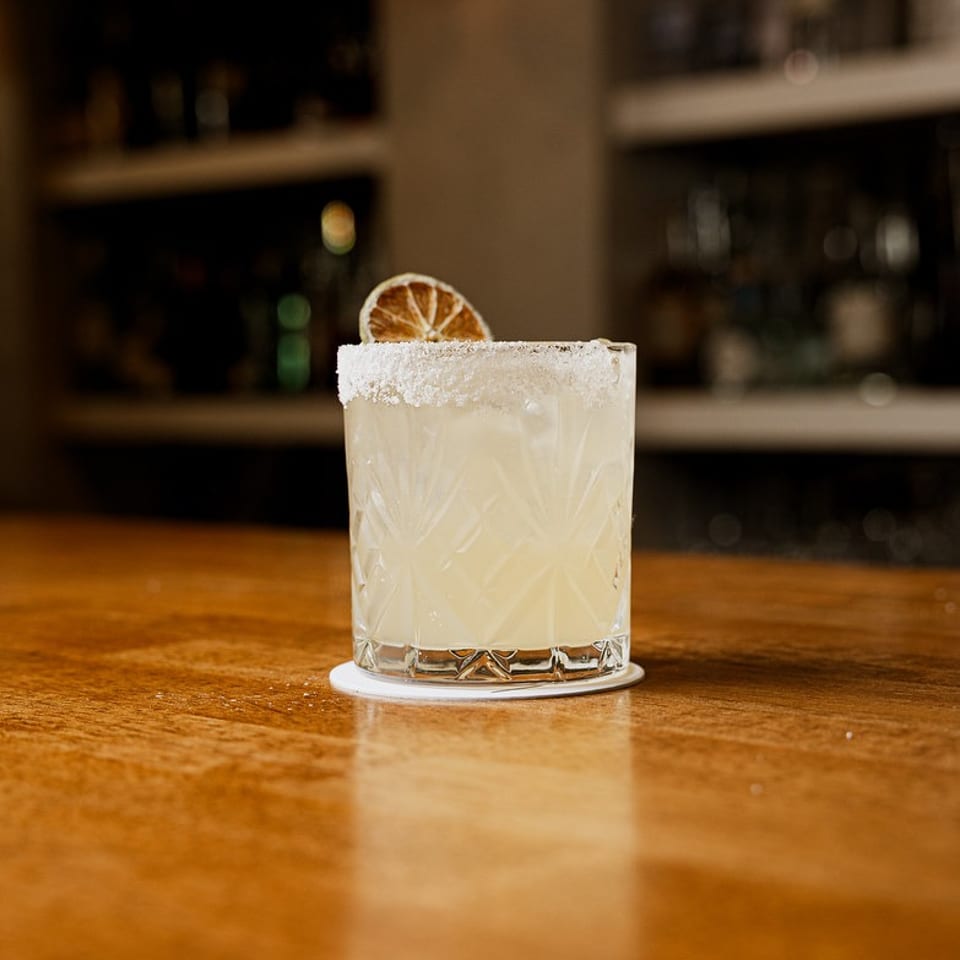 Light cocktail with a salted rim and dried citrus slice on a wooden bar.