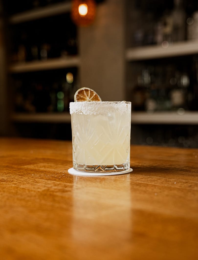 Light cocktail with a salted rim and dried citrus slice on a wooden bar.