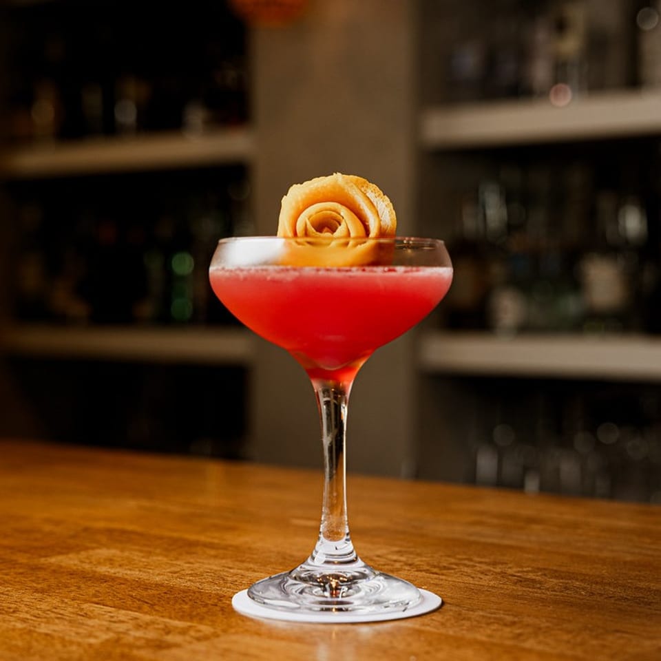 Red cocktail in a coupe glass with an orange peel rose garnish on a wooden bar.