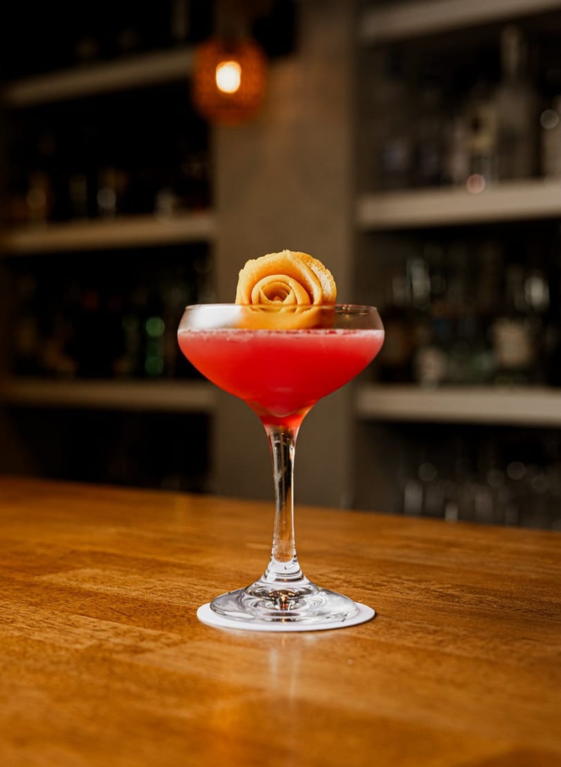 Red cocktail in a coupe glass with an orange peel rose garnish on a wooden bar.