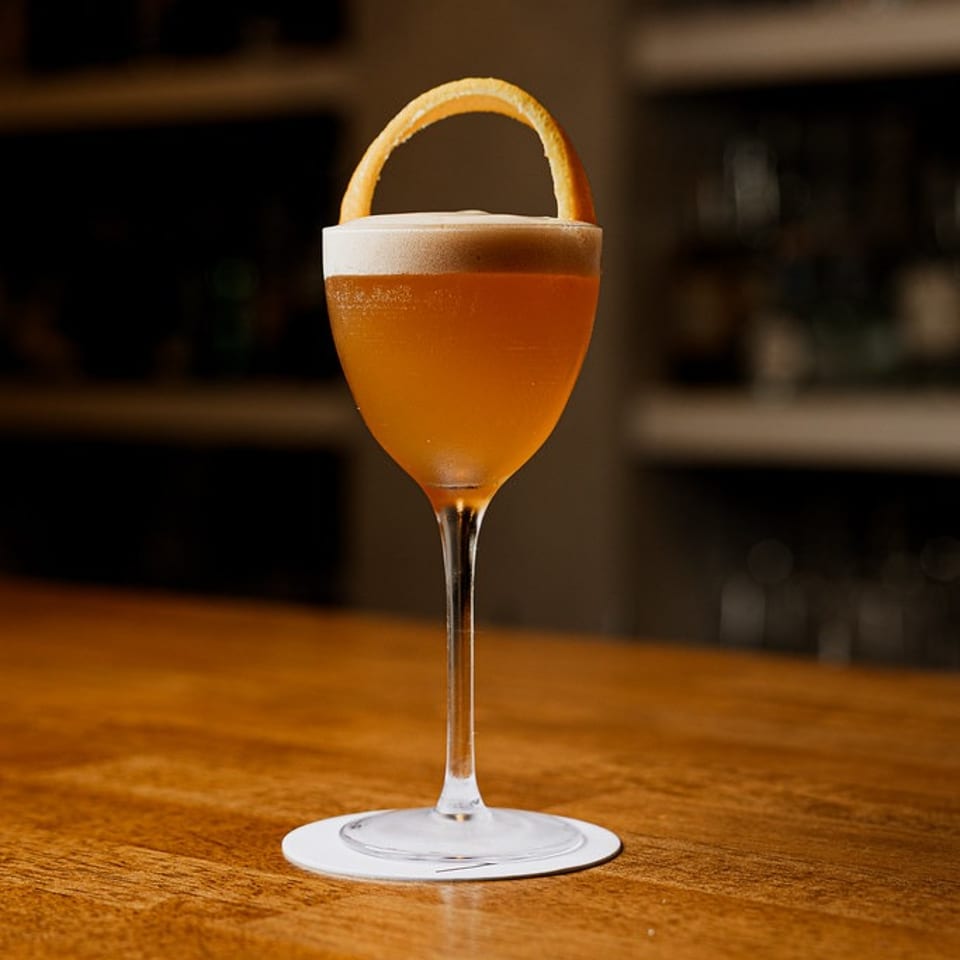 Elegant amber cocktail with foamy top and orange peel on a wooden bar.