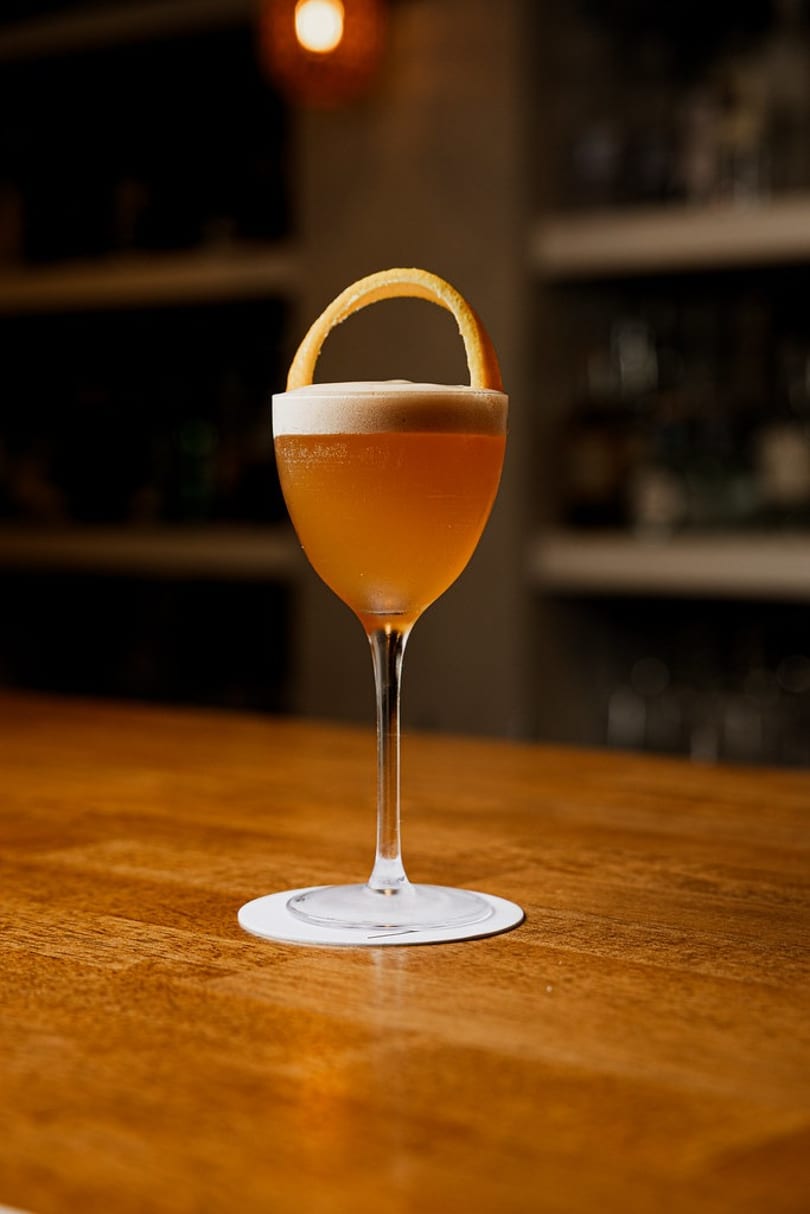 Elegant amber cocktail with foamy top and orange peel on a wooden bar.