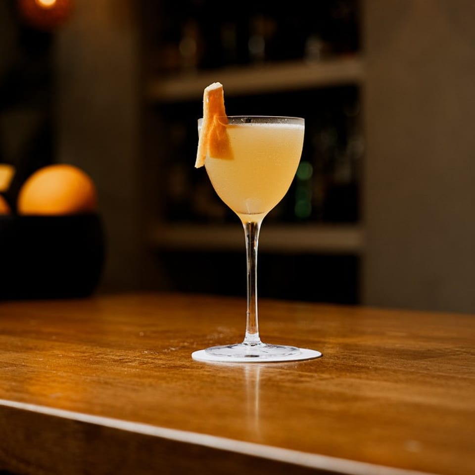 Elegant pale yellow cocktail with an orange peel garnish on a dark wooden bar.
