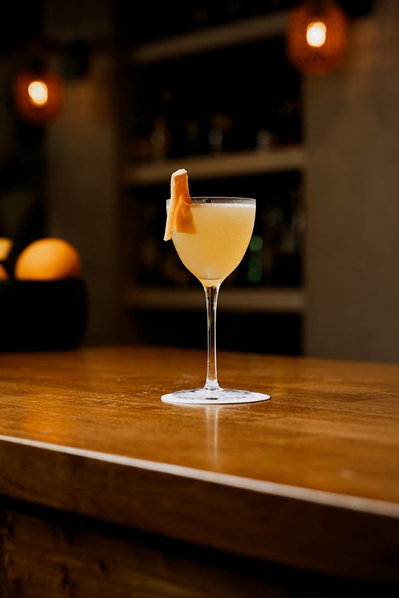 Elegant pale yellow cocktail with an orange peel garnish on a dark wooden bar.