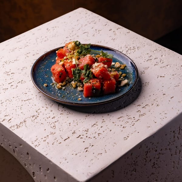 Diced watermelon salad with feta, herbs, and nuts on a blue plate, resting on a textured stone surface.