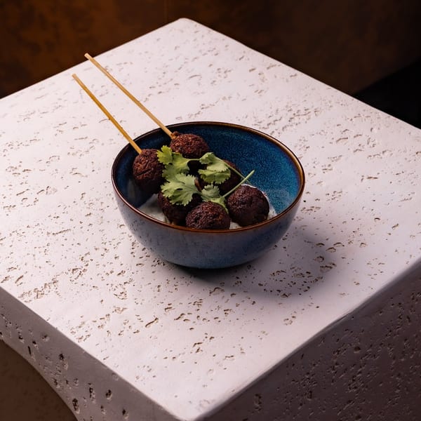 Dark, skewered appetizers with cilantro in a blue bowl on a textured stone table.