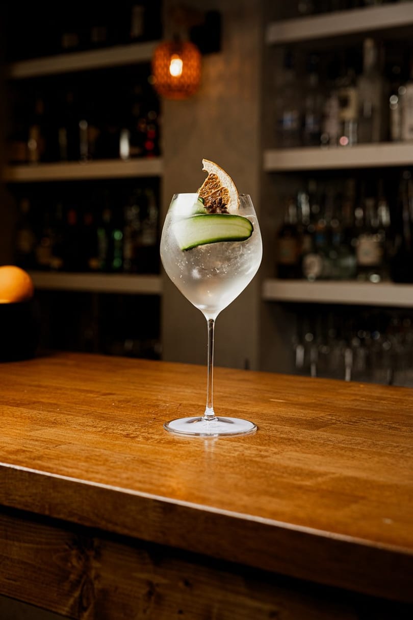 Refreshing cocktail with cucumber and dried orange slice on a polished wooden bar.