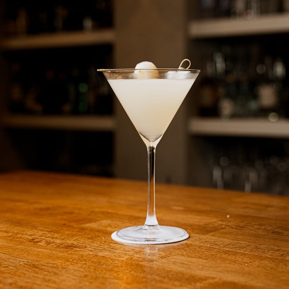 Light-colored martini cocktail with a lychee garnish on a polished wooden bar.