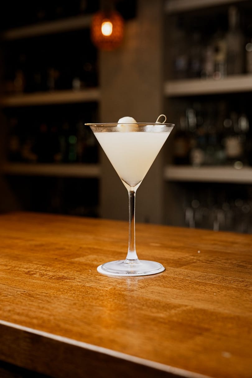 Light-colored martini cocktail with a lychee garnish on a polished wooden bar.