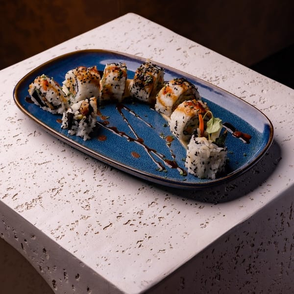Sushi rolls with sesame seeds and sauce on a blue plate, resting on a textured surface.
