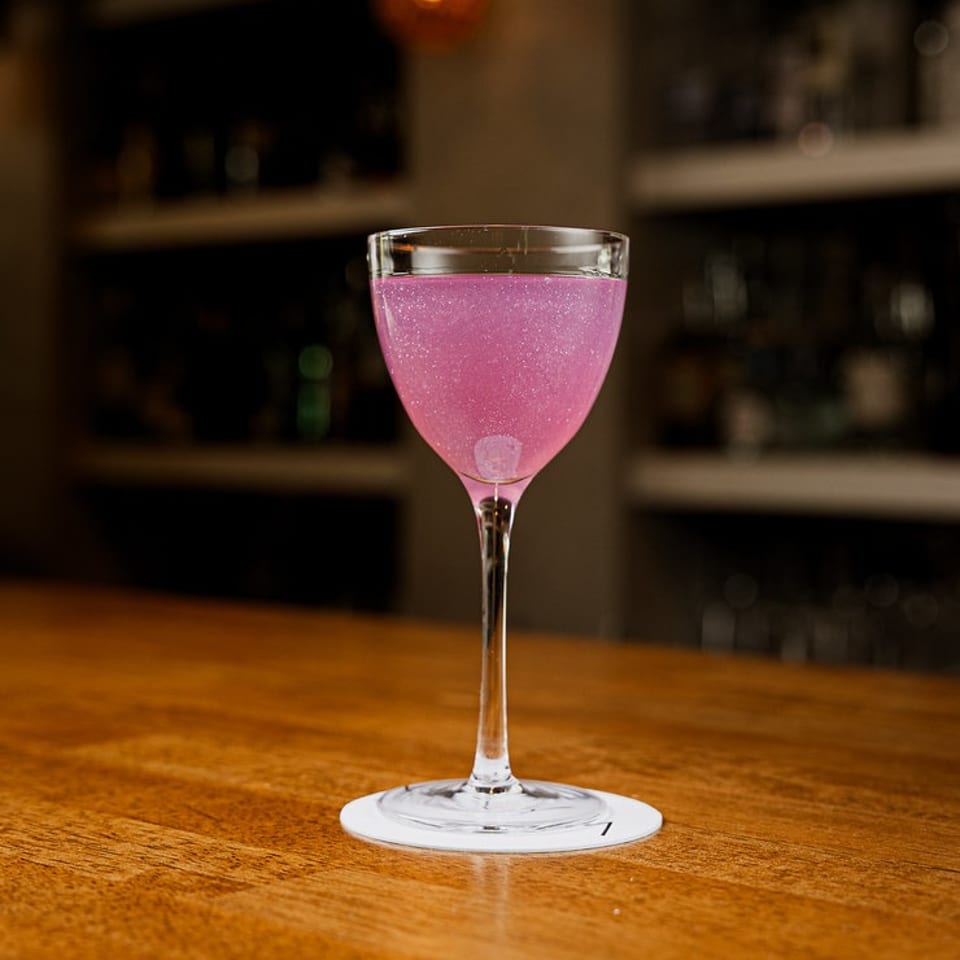 Vibrant pink sparkling cocktail in a coupe glass on a polished wooden bar.