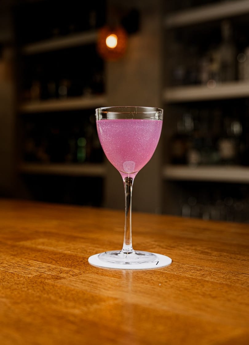 Vibrant pink sparkling cocktail in a coupe glass on a polished wooden bar.